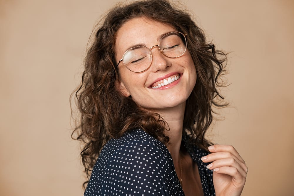 Young woman with curly hair and glasses smiling joyfully with eyes closed, showing off healthy teeth after dental care – dentist San Diego