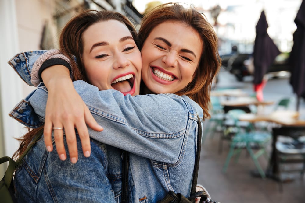 Two joyful young women in denim jackets hugging and laughing outdoors, showing off bright, beautiful smiles – dentist San Diego 