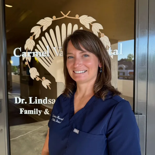 Dr. Lindsay Bancroft, DDS smiling and wearing navy scrubs in front of the front door of her office - Carmel Valley Dental Dr. Lindsay Bancroft, DDS smiling and wearing navy scrubs in front of the front door of her office - Carmel Valley Dental