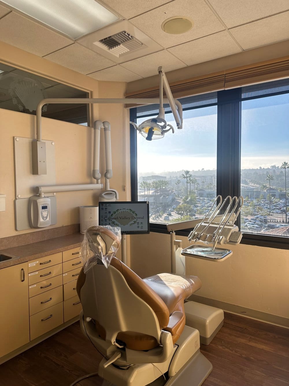 A modern dental exam room with a dentist's chair, equipment, and large windows offering a view of the outside - Carmel Valley Dental