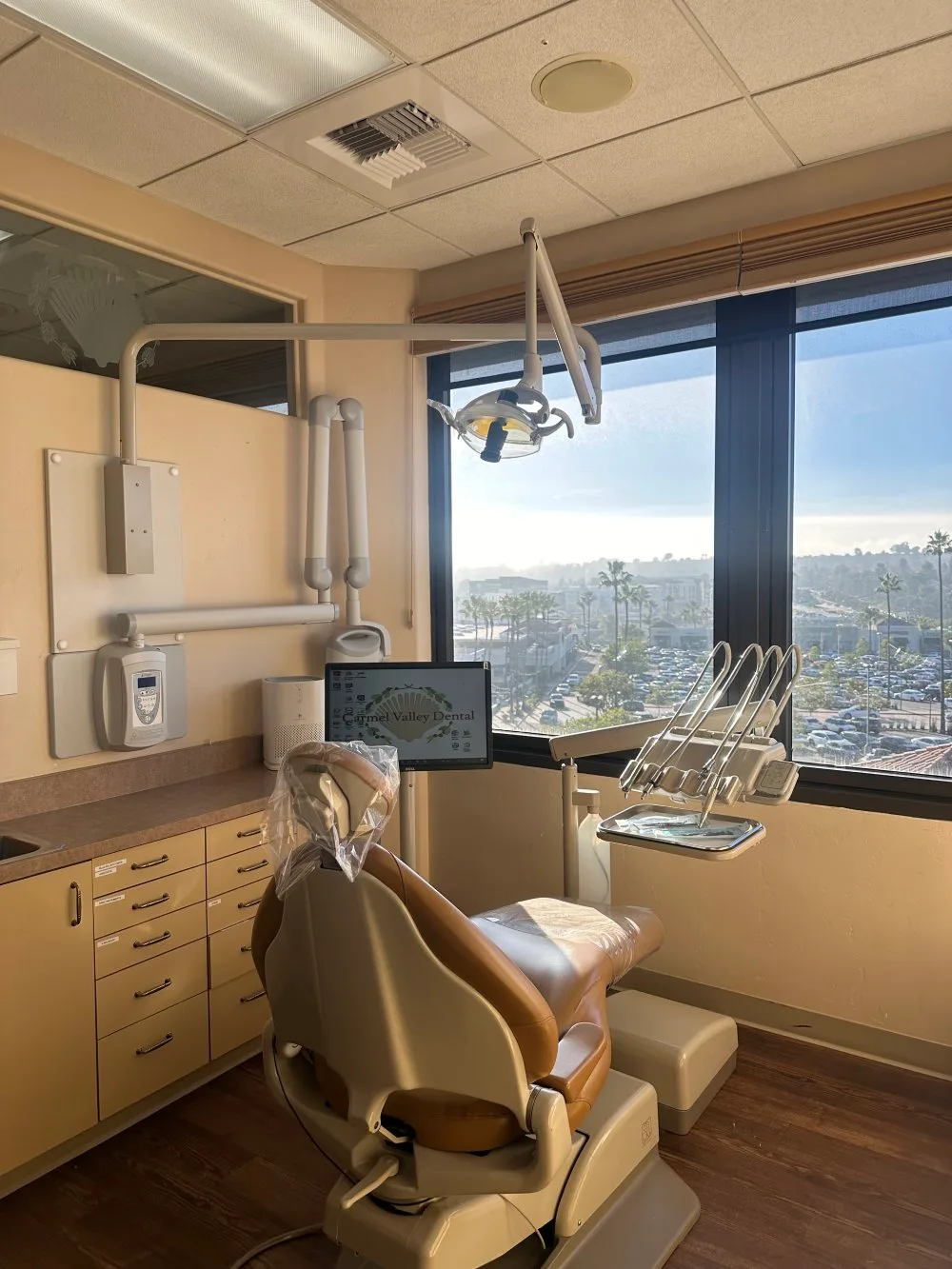 A modern dental exam room with a dentist's chair, equipment, and large windows offering a view of the outside - Carmel Valley Dental