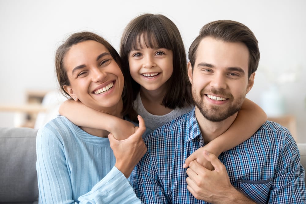 Family picture of smiling young mom and dad sit on couch posing with cute little daughter - Dental Cleaning