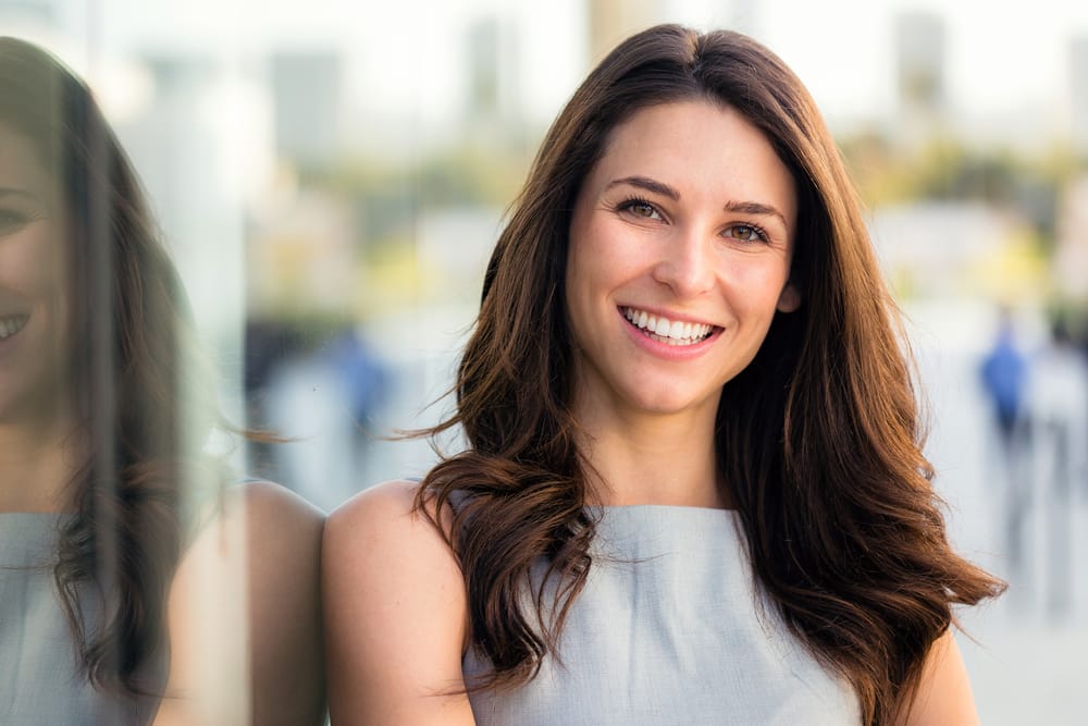 Confident Smile Following Emergency Dental Care – emergency dentist San Diego Young woman with long brown hair smiling brightly in an outdoor urban setting, reflecting restored dental health after emergency treatment – emergency dentist San Diego