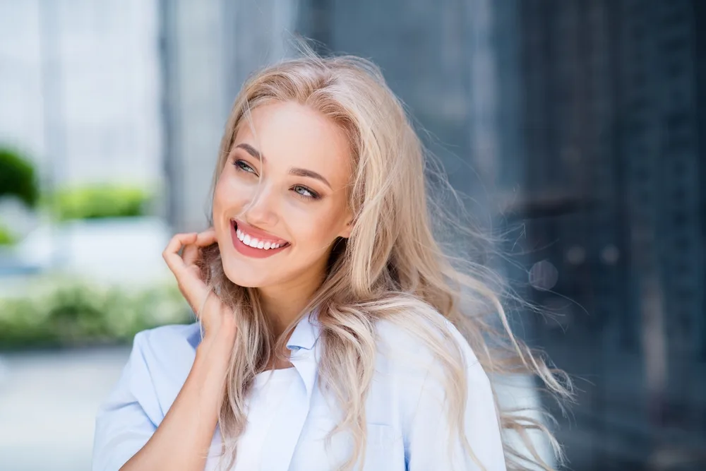 Confident Smile After Emergency Dental Care – emergency dentist San Diego Blonde woman smiling confidently outdoors, with her hand gently near her cheek, symbolizing restored comfort and dental wellness – emergency dentist San Diego