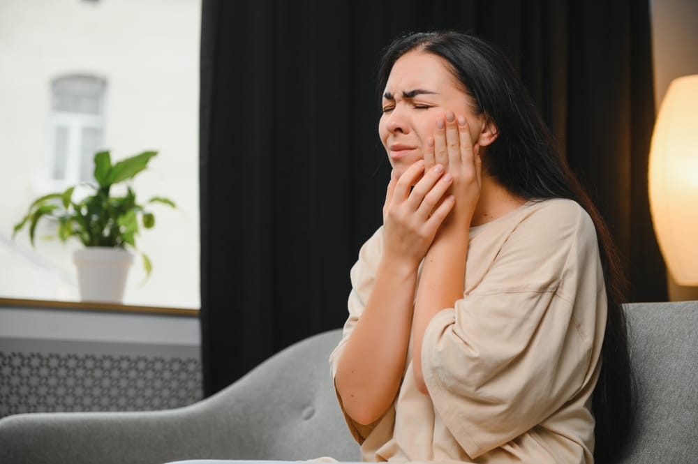 Woman Suffering from Sudden Tooth Pain – emergency dentist San Diego Young woman clutching her cheek in visible pain while seated on a couch, illustrating the need for urgent dental care – emergency dentist San Diego