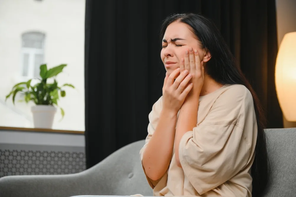 Woman Suffering from Sudden Tooth Pain – emergency dentist San Diego Young woman clutching her cheek in visible pain while seated on a couch, illustrating the need for urgent dental care – emergency dentist San Diego
