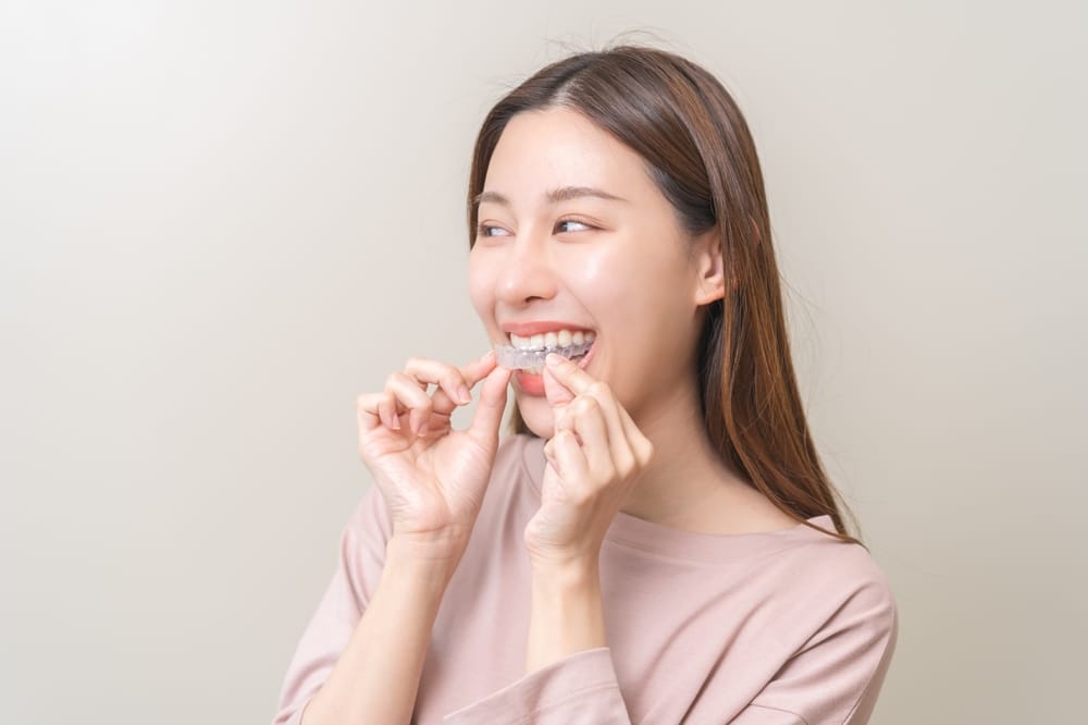 Young woman smiling while inserting a clear aligner onto her teeth, demonstrating orthodontic treatment options like Invisalign – dentist San Diego
