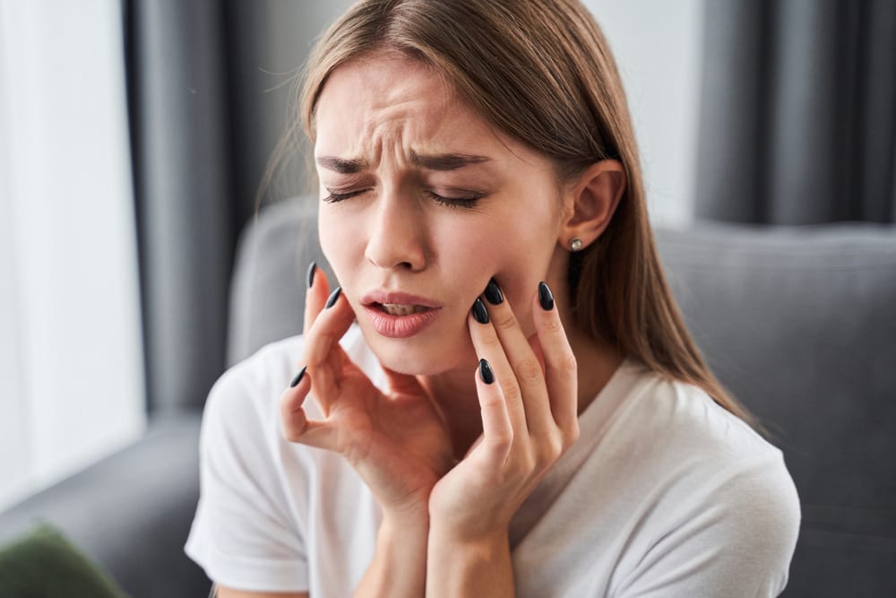 Frustrated woman holding both sides of her jaw in pain, possibly suffering from TMJ or toothache while seated at home – dentist San Diego 
